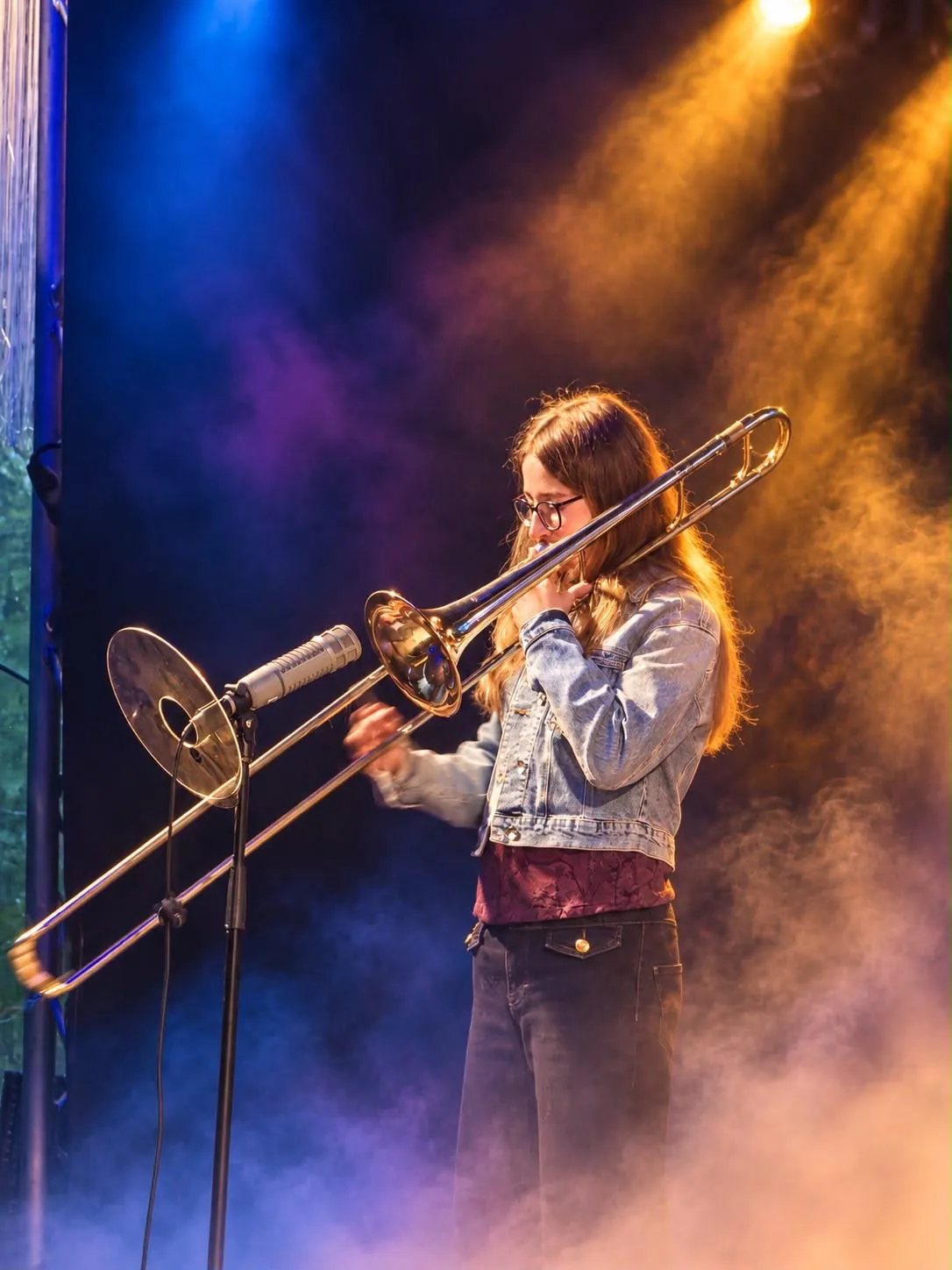 Luna Spiegel toca el trombón en el escenario, iluminada por focos azules y dorados entre el humo escénico.